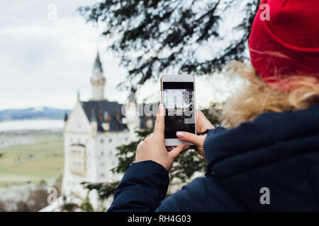 SCHWANGAU, Deutschland - Dezember 2018: Woman in Red hat ein Bild von Schloss Neuschwanstein auf Handy. Verschwommenen Hintergrund. Stockfoto