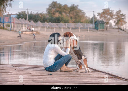 Porträt der schöne junge Frau sitzt am Fluss und umarmte ihre Hund Basset Hound. Glückliche Frau mag Zeit mit Ihrem Hund zu verbringen. Stockfoto