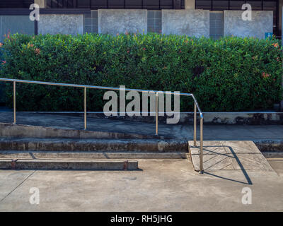 Alten grunge Rollstuhlrampe mit Metall Bar in der Nähe der leeren Parkplatz vor der WC-Gebäude mit grünen Bush Hintergrund. Stockfoto