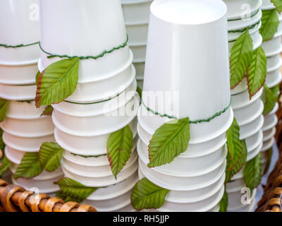 Weiß Einwegbecher mit der Oberseite nach unten. Stapel von White Paper cups Dekoration mit grünen Blättern im Warenkorb. Stockfoto