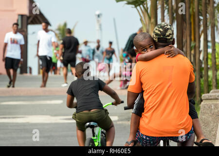 Durban, Südafrika - 6. Januar 2019: Ein schwarzer Mann ist mit dem Fahrrad mit seinem Kind in seine Arme draußen am Strand von Durban, Südafrika Stockfoto