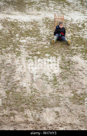 Junge stoppte am Hang, mit dem Schlitten sitzen auf einem Hügel mit sehr wenig Schnee Stockfoto