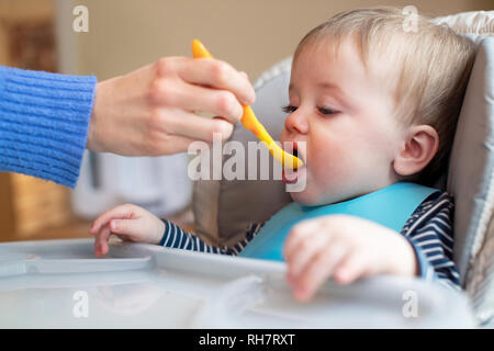 Baby Boy zu Hause in Hochstuhl Zugeführtes feste Nahrung von der Mutter mit Löffel Stockfoto