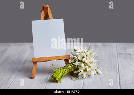 Blumenstrauß aus schneeglöckchen und eine kleine Staffelei mit einem weißen Papier und mini Gläser auf einem blauen Hintergrund. Grußkarte mit einem Platz für Text Stockfoto