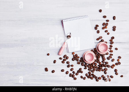 Zwei kleine rosa Tassen auf dem Tisch mit einem Löffel, Notepad und verstreute Kaffeebohnen auf weißem Holz- Hintergrund. Einladung oder Grußkarte. Ansicht von oben. Stockfoto