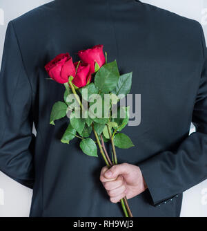 Eleganten Mann mit roten Rosen in der Hand hinter seinem Rücken. Überrascht Geschenk für Frau. Romantisch und Valentines Tag Urlaub Konzept. Nahaufnahme Stockfoto