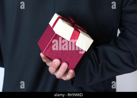 Mann in Anzug, rote Rose Geschenk Box hinter seinem Rücken. Überrascht Geschenk für Frau. Romantisch und Valentines Tag Urlaub Konzept. Nahaufnahme Stockfoto