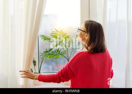 Frau schaut aus dem Fenster, und öffnet die Gardinen. Ansicht von der Rückseite. Stockfoto