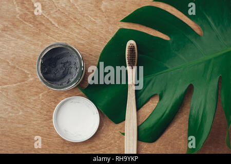 Natürliche Zahnpasta Aktivkohle und Bambus Zahnbürste auf Holz- Hintergrund mit grünen monstera Blatt. Kunststoff freie Schönheit essentials, Zähne kümmern. Stockfoto
