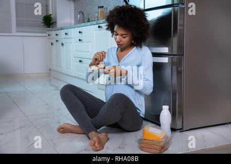 Junge schwangere Frau Essen Stück Kuchen sitzen vor dem offenen Kühlschrank in der Küche Stockfoto