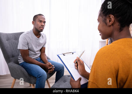 Junge afrikanische Mann sitzt auf Stuhl in der Nähe der Psychologin mit Zwischenablage Stockfoto