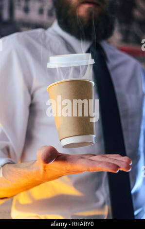 Schwebend in der Luft Papier Tasse mit heißem Kaffee. Barista, ein bärtiger junger Mann in einem weißen Hemd mit Krawatte, schafft Wunder - wirbt mit seinem Drink, Ca Stockfoto