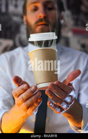 Schwebend in der Luft Papier Tasse mit heißem Kaffee. Barista, ein bärtiger junger Mann in einem weißen Hemd mit Krawatte, schafft Wunder - wirbt mit seinem Drink, Ca Stockfoto