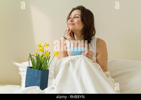 Erwachsene Frau im Bett zu Hause, morgen erhalten eine Überraschung Blumenstrauß. Liest Karte und glücklich Stockfoto