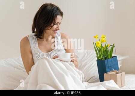Frau mittleren Alters mit Geschenk, Blumenstrauß gefreut, morgen im Bett bei einer Tasse Kaffee. Gefühl von Glück, Freude, Überraschung. Stockfoto