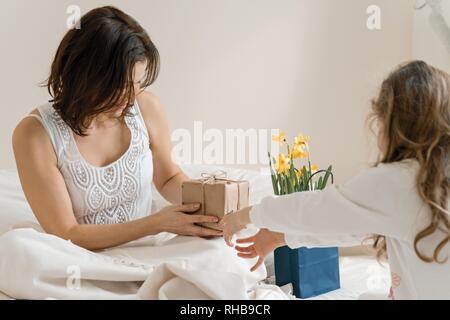 Muttertag. Kleine Tochter hält Geschenk und Blumen für Ihre Mutter. Hintergrund Interieur der Zimmer, der Mutter im Bett, morgen. Stockfoto