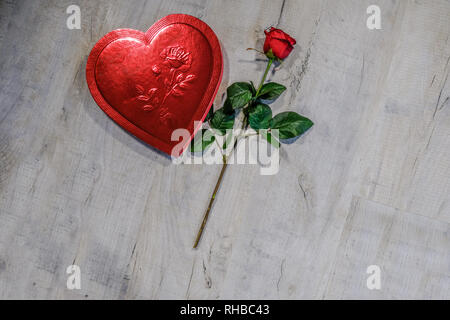 Rotes Herz Form Box mit einer roten Rose neben liegen. Auf grauem Hintergrund mit Holz copyspace platziert. Stockfoto