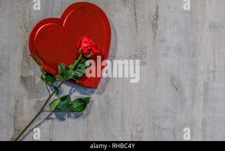 Rotes Herz Form Box mit einer roten Rose lyingon oben auf der Box. Auf grauem Hintergrund mit Holz copyspace platziert. Stockfoto