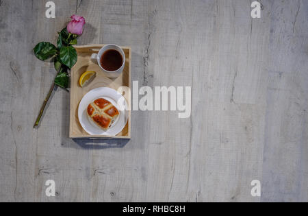Flachbett Top Shot eines Fachs mit Zitrone Tee- und Hot Cross bun. Eine schöne Single rosa Rose Stammzellen neben liegen. Auf einem Dritten mit Copyspace besi gelegen Stockfoto