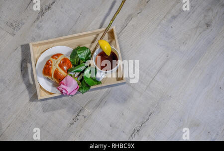 Flachbett Top Shot eines Fachs mit Zitrone Tee- und Hot Cross bun. Eine schöne Single rosa Rose Stammzellen neben liegen. Auf einem Dritten schräg gelegen. Stockfoto