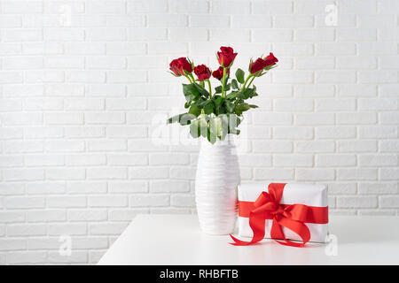 Rote Rosen, weiße Vase neben einer Geschenkbox mit roter Schleife gegen Weiße brocks Wand Stockfoto