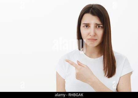 Düster, traurig und verzweifelt cute brunette Mädchen im weißen T-Shirt mit gerunzelter Stirn, schürzte die Lippen und zeigte mit Eifersucht oder bedauern Links, über unlautere Stockfoto