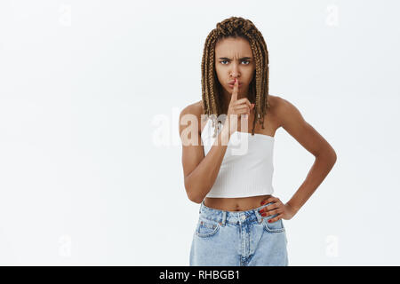 Shh Ich denke hier. Ernsthaft gestört und herrisch dunkelhäutige Frau mit Dreadlocks stirnrunzelnd shushing an Kamera mit Index Finger über den Mund, Stockfoto