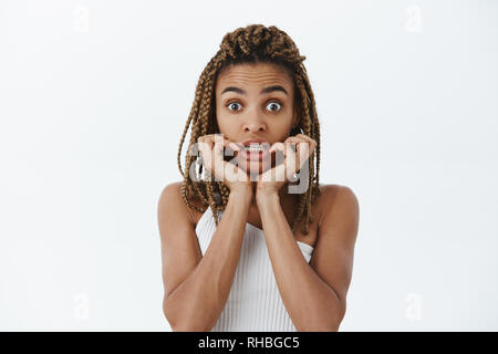 Mädchen reagieren auf besorgniserregende Nachrichten schockiert. Portrait von überrascht und erstaunt, sprachlos dunkelhäutige Frau mit Dreadlocks Aufeinanderpressen der Zähne die Erntehaltefinger Stockfoto