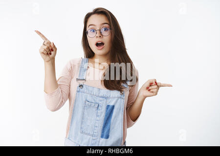 Neugierig und intelligente junge Kreative 20 s Europäische Weiblich in Gläsern und Denim Overalls offenen Mund in Frage gestellt und beeindruckt zeigen seitwärts fasziniert Stockfoto