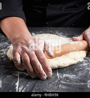 Koch in einem schwarze Tunika, rollt Teig für Pizza auf einem Schwarzen Tisch mit einem hölzernen Rolling Pin Stockfoto