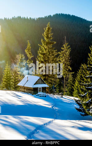 Ferienhaus aus Holz auf einem schneebedeckten Rasen in die Berge. Die Tannen sind mit morgen Strahlen behandelt. Rauch aus dem Kamin des Hauses. Winter i Stockfoto