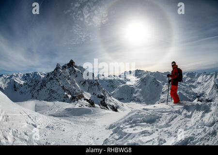 Eine männliche Skifahrer steht auf dem Grat zwischen den Skigebieten von Courchevel und Meribel in Frankreich mit einer Sonne halo durch Eiskristalle verursacht in der Luft hinter sich. Stockfoto