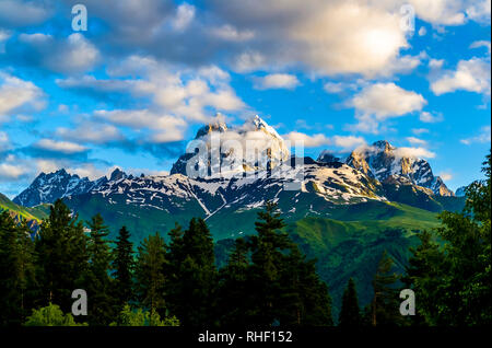 Hohen schneebedeckten Gipfel auf einem Hintergrund von weißen Wolken und blauer Himmel. Morgen scheint die Sonne an der Wand. Grüne Hügel und dichten Wälder auf der Piste. Mount Stockfoto