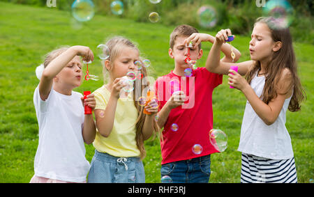 Gerne Jungen und Mädchen im Grundschulalter stehen auf grünem Gras und Seifenblasen Stockfoto