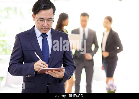 Portrait von asiatischen Geschäftsmanns, der Verwendung von smart Tablet mit Business Team im Hintergrund bei Office Fenster mit für Coporate arbeiten Hintergrund Stockfoto