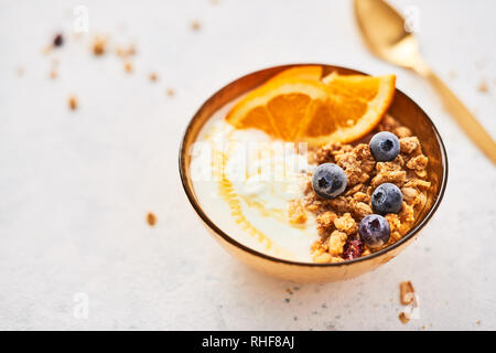 Gesundes Frühstück. Schüssel mit hausgemachtem Müsli mit Joghurt, Honig, frischen Orange und gefrorene Heidelbeeren auf weißem Abstrakt Hintergrund. Golden Utensilien. Top Stockfoto