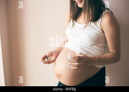 Seitenansicht der lächelnden schwangere Frau mit dicken Bauch zu Hause stehen und Vitamine mit Wasser. Junge zukünftige Mutter über Gesundheit interessieren. Konzept der Erwartung und der elterlichen Liebe. Stockfoto
