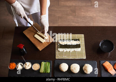 Detailansicht des Prozesses der Vorbereitung Rolling Sushi. Hände in Einweghandschuhe slicing Omelett auf Holzbrett Stockfoto