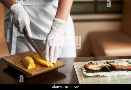 Detailansicht des Prozesses der Vorbereitung Rolling Sushi. Hände in Einweghandschuhe slicing Omelett auf Holzbrett Stockfoto