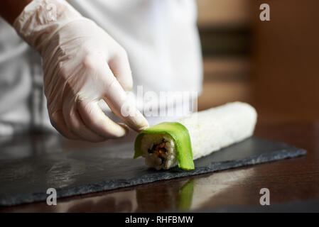 Nahaufnahme der Prozess der Vorbereitung Rolling Sushi an. Weibliche Hand in Hand ziert Roll mit in Scheiben geschnittenen Avocado Stockfoto