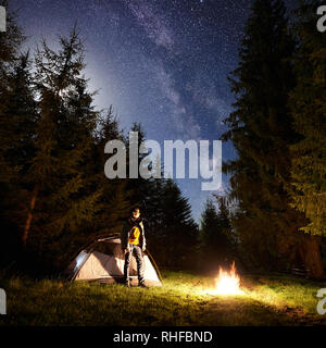 Campingplatz in der Nacht. Touristische Zelt auf Waldlichtung und junge Wanderer stand vor der brennenden Lagerfeuer unter dunklen blauen Sternenhimmel auf Pinien im Hintergrund. Schönheit der Natur und Tourismus Konzept. Stockfoto