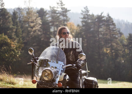 Schöne bärtige Motorradfahrer mit langen Haaren in schwarzer Lederjacke und Sonnenbrille sitzen auf Cruiser Motorrad auf unscharfen Hintergrund der grünen ruhigen ländlichen Landschaft und das Licht nebligen Himmel. Stockfoto