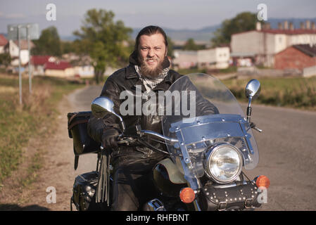 Portrait von Stattlichen glücklich lächelnde bärtigen Biker in schwarzem Leder Jacke sitzen auf modernen Motorrad auf Land am Straßenrand auf unscharfen Hintergrund der kleinen Stadt bauten auf sonnigen Sommertag. Stockfoto