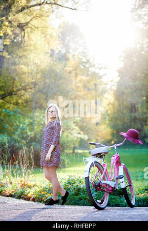 Gerne blonde modische Langhaarigen attraktive Frau im kurzen Kleid stehen an der Dame mit dem Fahrrad auf asphaltierten Sommer Park Alley auf schönen, grünen und goldenen Bäumen Hintergrund in den Abend. Stockfoto