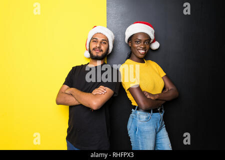 Junge glücklich Mixed Race Paar, Inder und afrikanische Frau in santa Hüte stehen auf gelb schwarz hintergrund Stockfoto