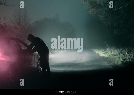 Ein einsamer hooded Abbildung in ein Auto suchen. Auf eine gespenstische Silhouetted, Land nebligen Winter Straße bei Nacht. Mit einem kalten, Körnigen bearbeiten. Stockfoto