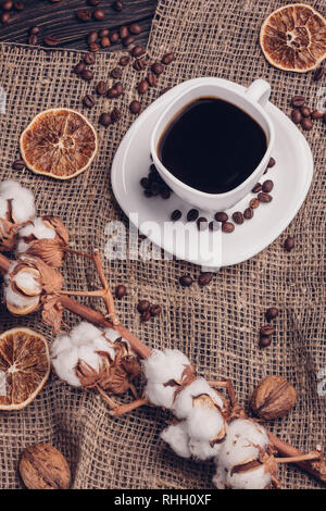 Schwarzer Kaffee in weisser Tasse mit Untertasse mit Kaffeebohnen, trockene Baumwolle auf einem hölzernen Hintergrund, mit Sackleinen abgedeckt Stockfoto