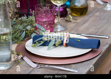 Blau serviette in Ring auf weiße Platte mit Kupfer Ladegerät, setzen Sie die Einstellung auf Holz Outdoor Party Esstisch mit Blumen, Glas, und Kerze. Stockfoto