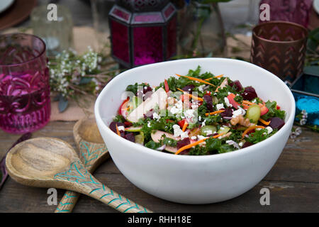 Große weiße Schale mit bunten gesund Grünkohl Salat mit Holz-, Löffel auf Outdoor Party Tisch mit lila Laterne, Gläser, und Kerzen. Stockfoto