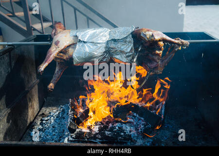 Gebackene geschlachtete Schweine am Spieß kochen auf einem großen handgefertigte Röster über dem offenen Feuer. In Folie eingewickelt. Gebäude auf einem Hintergrund. Stockfoto